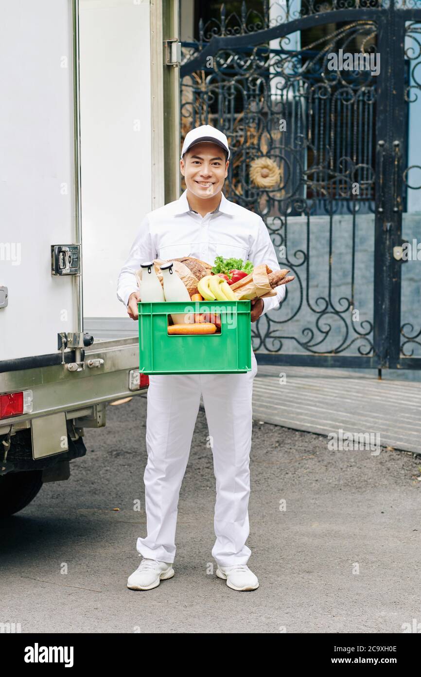 Portrait eines gutaussehenden jungen vietnamesischen Kuriers, der eine Plastikkiste mit Milchflaschen, frischem Brot und Obst hält Stockfoto