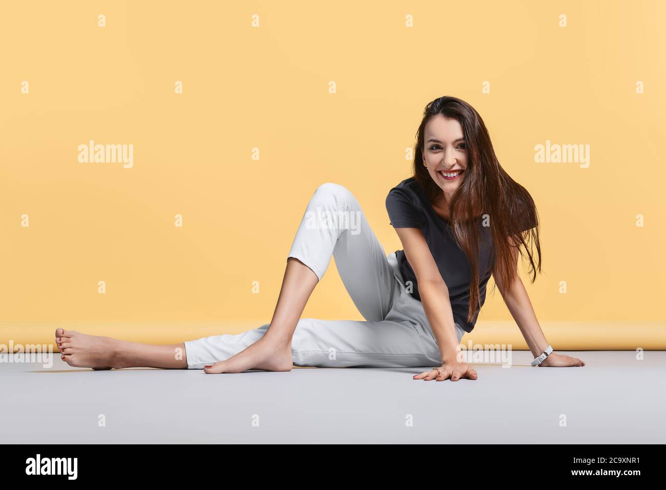 Happy barfuß junge Frau in lässigen Hosen und Hemd sitzt auf dem Boden im Studio auf gelbem Hintergrund Stockfoto