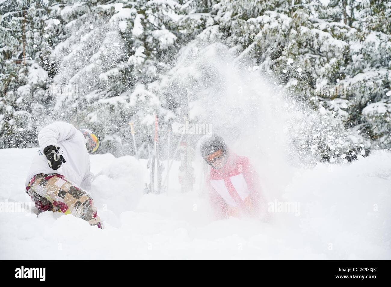 Junge Paar Skifahrer in Skianzügen und Helmen werfen frischen Pulverschnee in die Luft und lächeln. Mann und Frau in Skibrille gute Zeit im Skigebiet mit schönen verschneiten Bäumen auf dem Hintergrund. Stockfoto