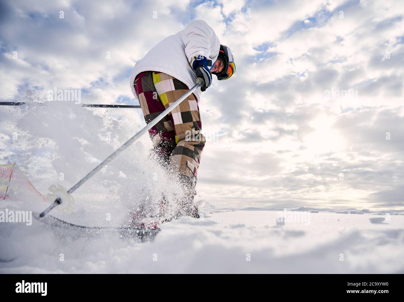 Low-Angle-Ansicht Momentaufnahme der Beine des Skifahrers machen einen Sprung auf weißen schneebedeckten Oberfläche gegen schönen bewölkten Himmel. Mann trägt bunte Skihose und weiße Winterjacke. Konzept der Wintersport-Aktivitäten Stockfoto