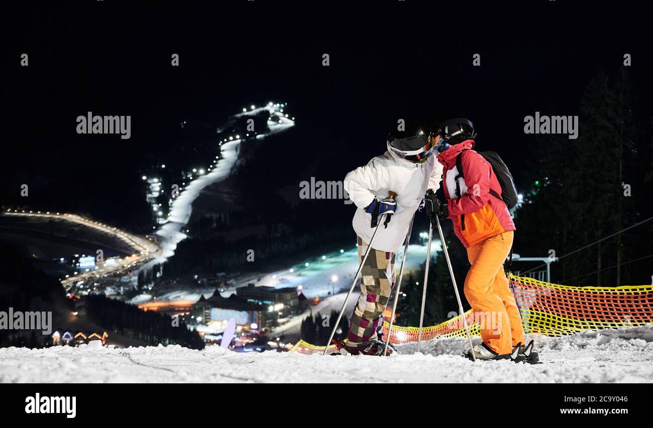 Ein paar Touristen stehen auf dem Hügel küssen vor erstaunlichen Nacht Hintergrund der beleuchteten Skipisten, Kopierer Raum. Konzept des aktiven Lebensstils, Nachtskifahren und Beziehungen. Stockfoto