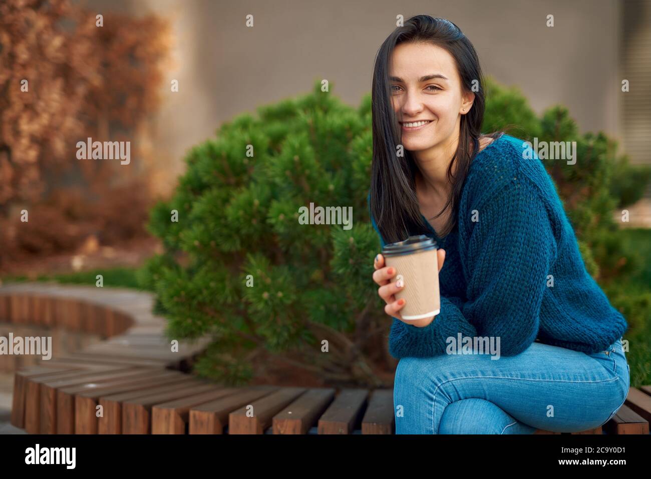 Charmantes junges Model posiert auf einem Holzsitz in einem Park, lehnt sich mit Ellbogen auf ihr Knie, lächelt und schaut in die Kamera Stockfoto