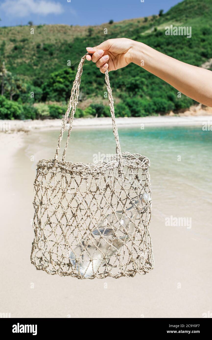 Frau hält Stroh Weidenwiederverwendbare Tasche mit Glasflasche für Wasser. Plastik frei und Zero Waste Lifestyle. Weißer Sandstrand mit klarem türkisfarbenem Wasser im Hintergrund. Stockfoto