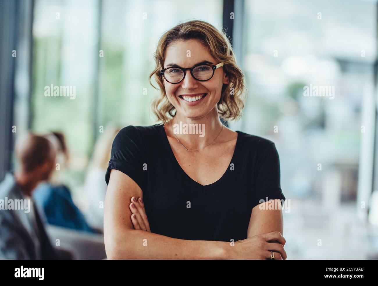 Porträt einer erfolgreichen professionellen Frau mit gekreuzten Armen. Lächelnde Geschäftsfrau, die im Büro steht und die Kamera anschaut. Stockfoto