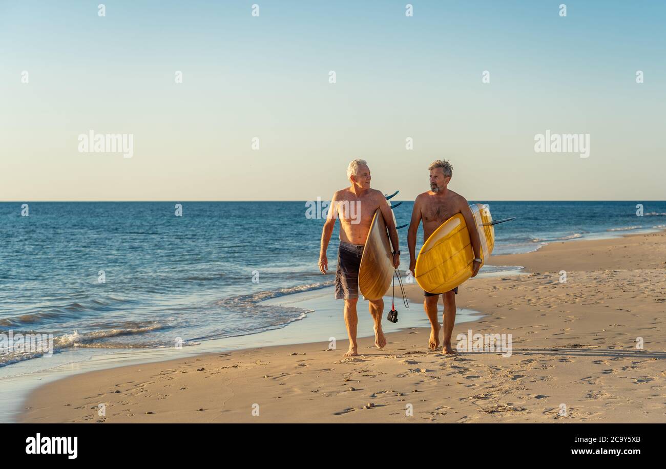 Zwei reife Männer, die mit Surfbrettern am wunderschönen Strand spazieren und das Paradies und den Lebensstil im Ruhestand genießen. Attraktiv fit ältere Erwachsene Freunde Spaß haben Stockfoto