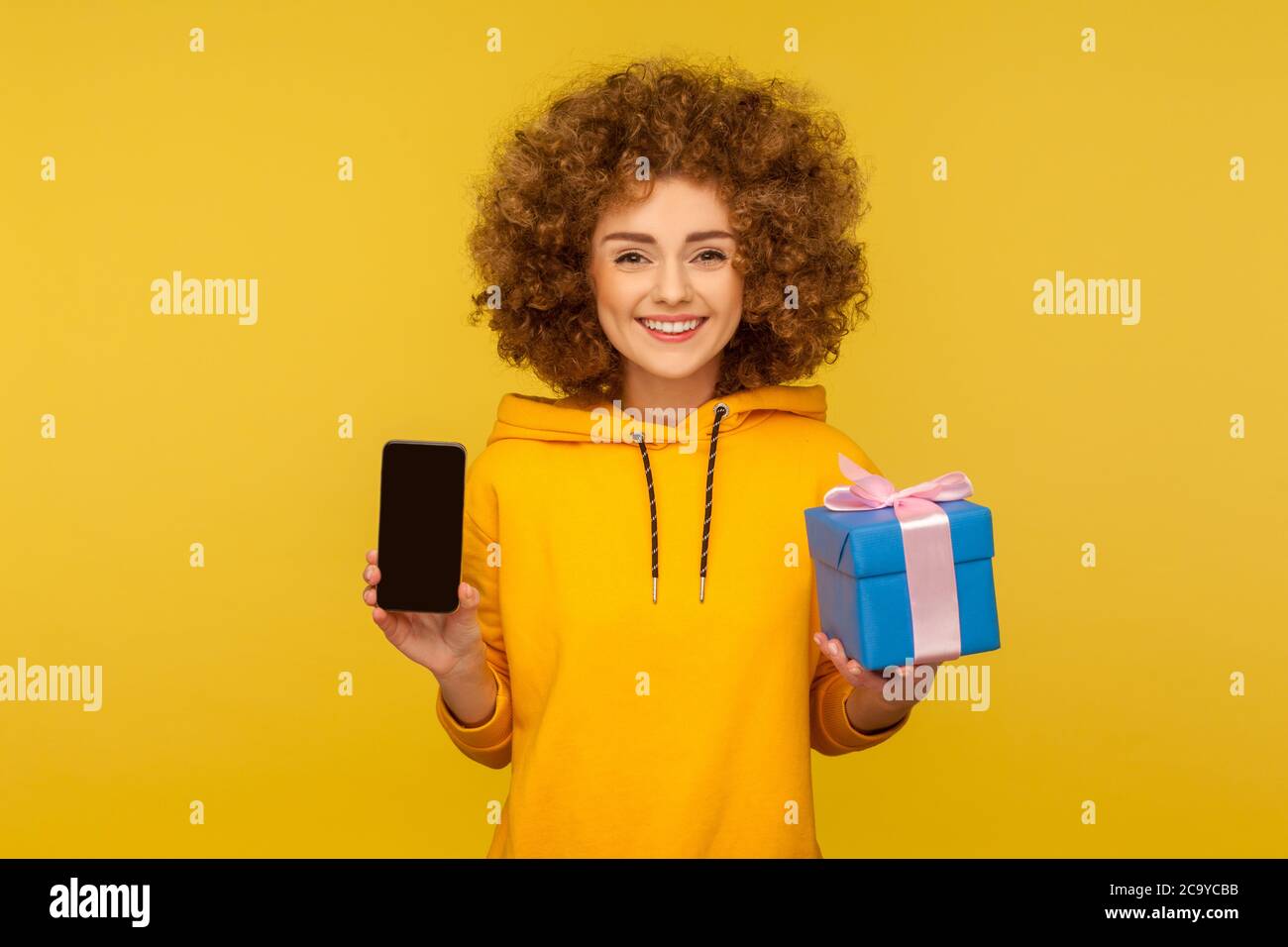 Einkaufen, Geschenke online kaufen. Porträt von glücklichen lockigen jungen Frau im urbanen Stil Kapuzenpullover mit Handy und Geschenkbox, lächelnd vor der Kamera. Stockfoto