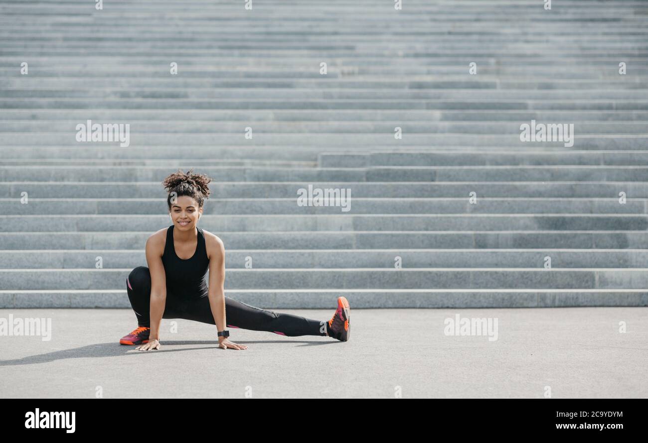 Erschwinglicher Sport im Freien. Lächelnde afroamerikanische Frau in Sportkleidung mit Fitness-Tracker und Kopfhörer tun Stretch für Bein, in der Nähe der Stadt Treppe Stockfoto