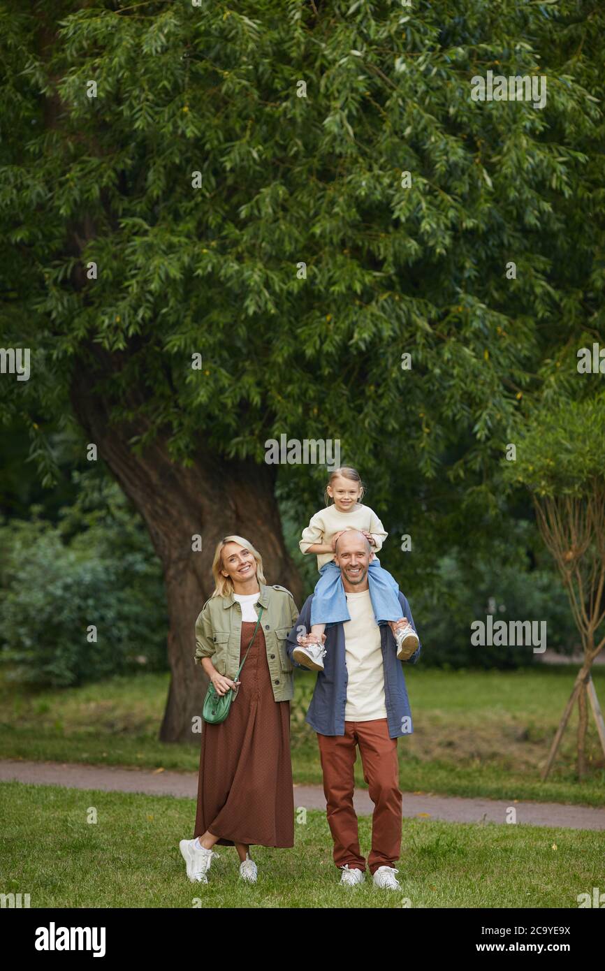 Vertikale Weitwinkel-Porträt von glücklichen modernen Familie zu Fuß im Park zusammen mit niedlichen kleinen Mädchen auf Väter Schultern sitzen, alle Blick auf die Kamera Stockfoto