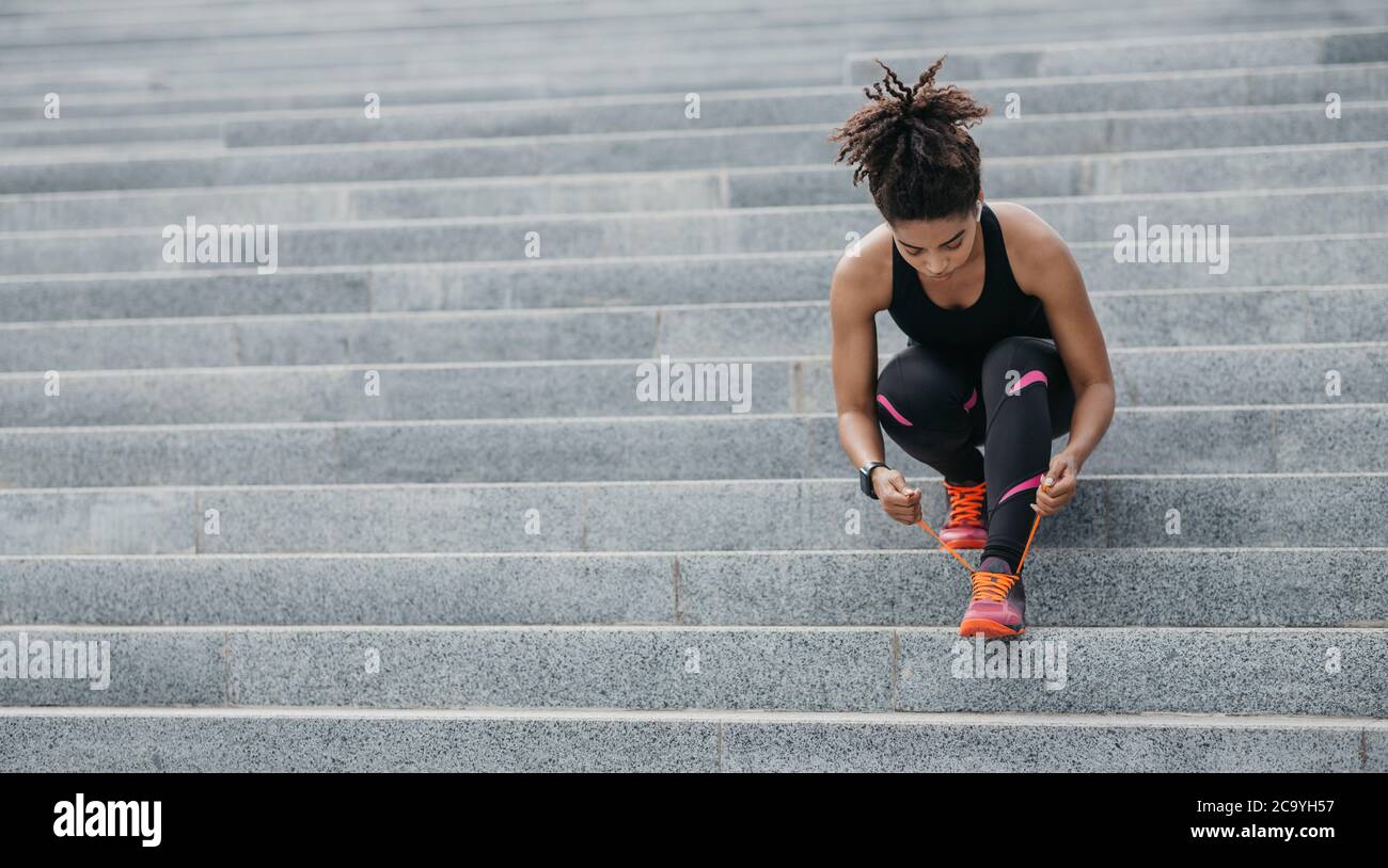 Muskulöse afroamerikanische Frau in Sportbekleidung, mit modernen Kopfhörern, Fitness-Tracker, Schnürsenkel auf Turnschuhen auf Treppen binden Stockfoto