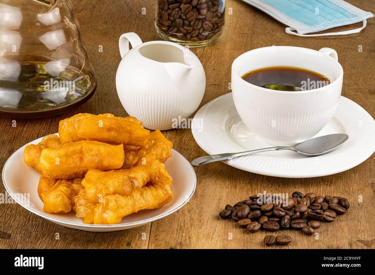 Frühstück mit frittiertem Teigstab in einer weißen Keramikplatte und schwarzem Kaffee in einer weißen Keramikschale mit einem Stapel Kaffeebohnen auf einem Holztisch. Stockfoto
