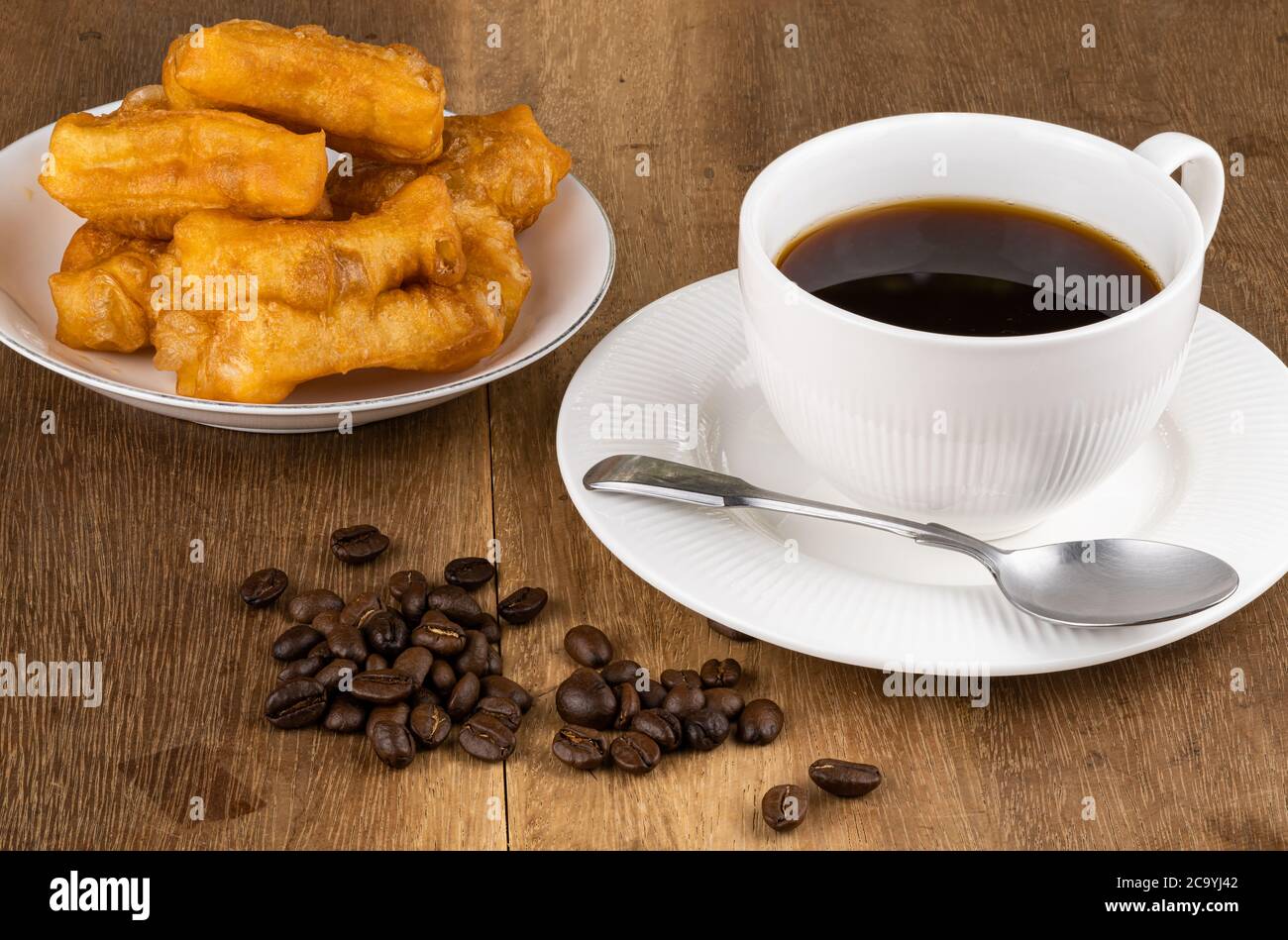 Normales Frühstück mit frittierten Teig Stick in einer weißen Keramikplatte, Stapel von Kaffeebohnen heißen schwarzen Kaffee in einer weißen Keramik-Tasse und Metalllöffel auf Stockfoto