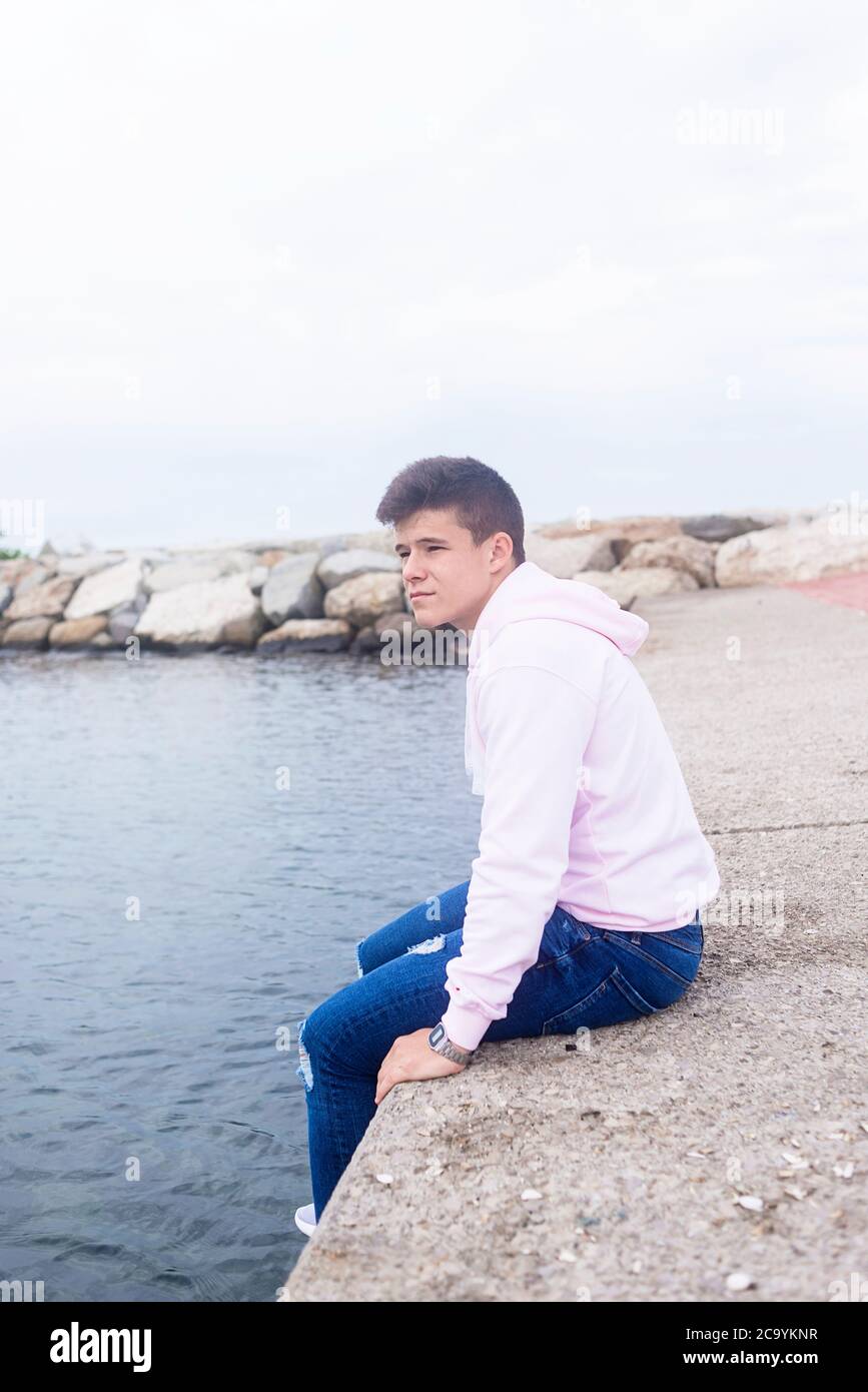 Junger nachdenklicher Teenager, der auf Wellenbrechern sitzt, während er die Kamera anschaut Stockfoto