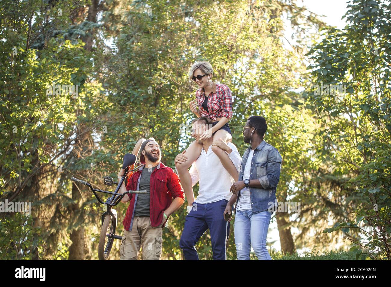 Multi-ethnische Gruppe von Menschen gehen mit ihrem männlichen Freund zu bmx Wettbewerb in der städtischen Sommerpark, Junge fröhliche Mann geben Huckepack Fahrt zu seinem blo Stockfoto