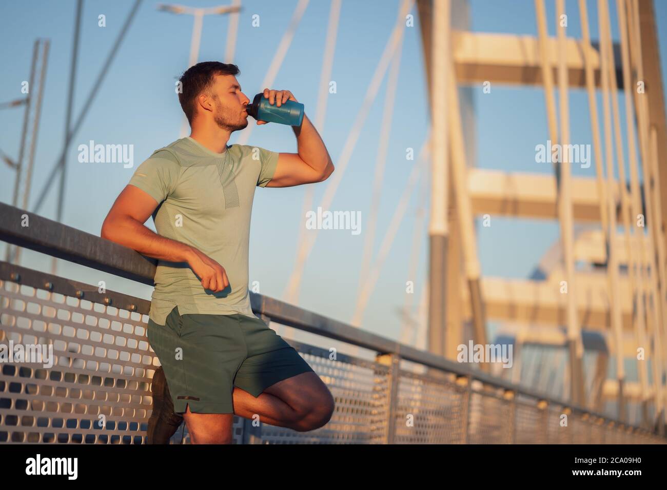 Der junge Mann trainiert draußen auf der Brücke in der Stadt. Er trinkt Wasser. Stockfoto