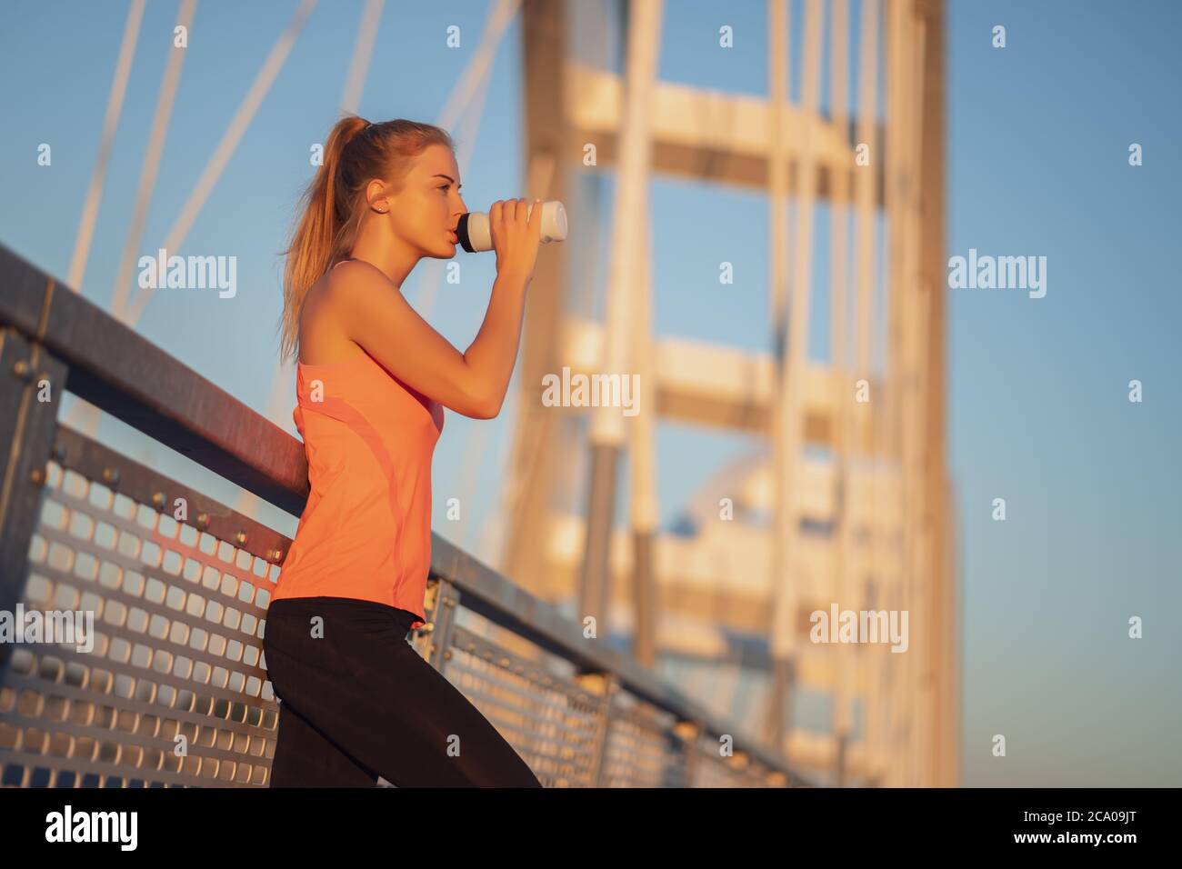 Junge Frau trainiert draußen auf der Brücke in der Stadt. Sie trinkt Wasser. Stockfoto