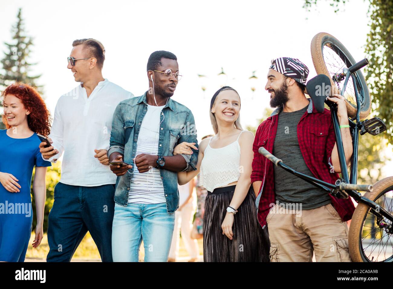 Multi-ethnische Gruppe von Menschen gehen mit ihrem männlichen Freund zu bmx Wettbewerb in der städtischen Sommerpark, Junge fröhliche Mann geben Huckepack Fahrt zu seinem blo Stockfoto
