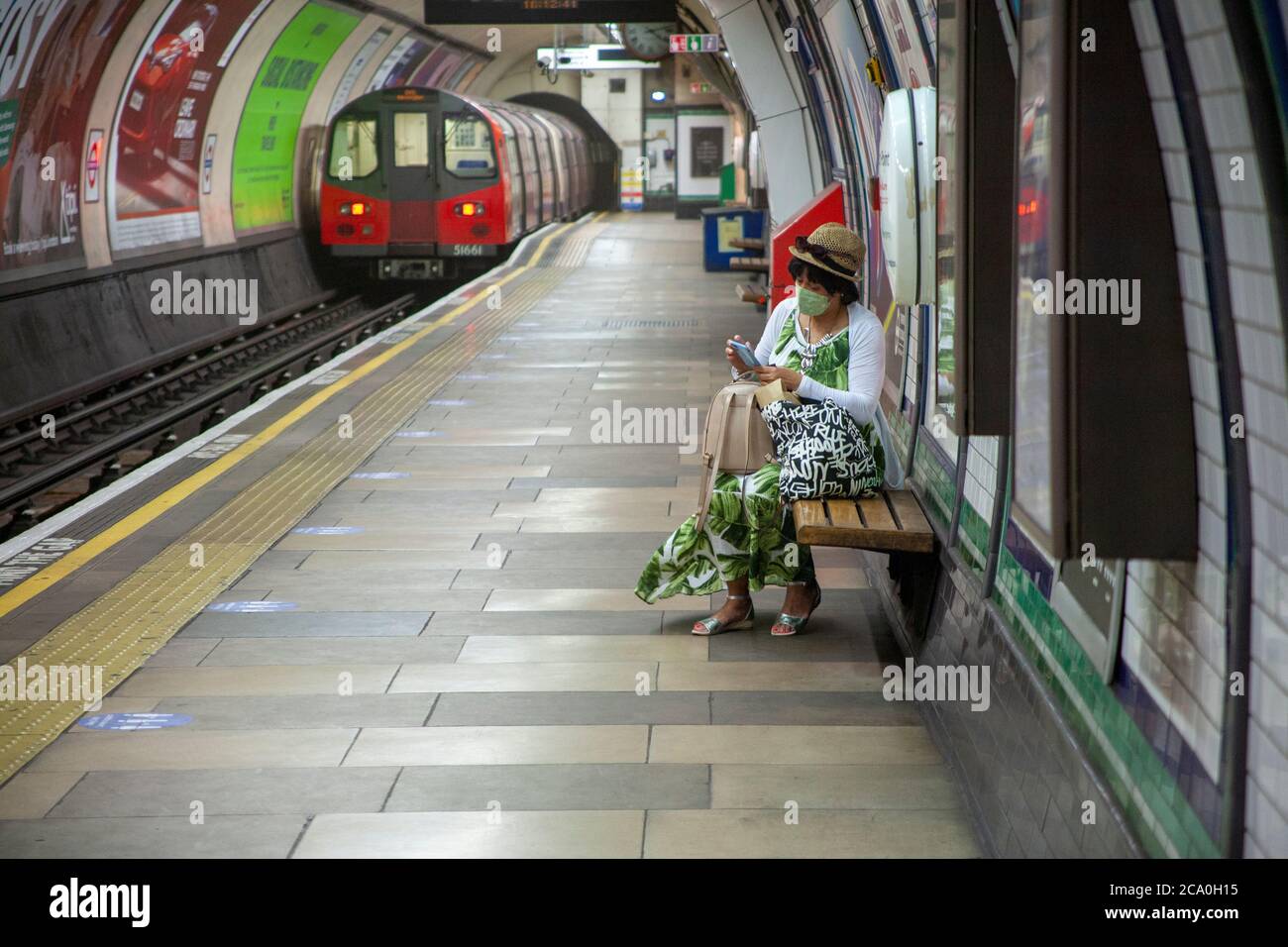 Frau mittleren Alters, trägt eine Gesichtsbedeckung, Hut und ein Sommerkleid, warten auf eine U-Bahn-Zug auf einem leeren, London Underground, Plattform. London, England Stockfoto