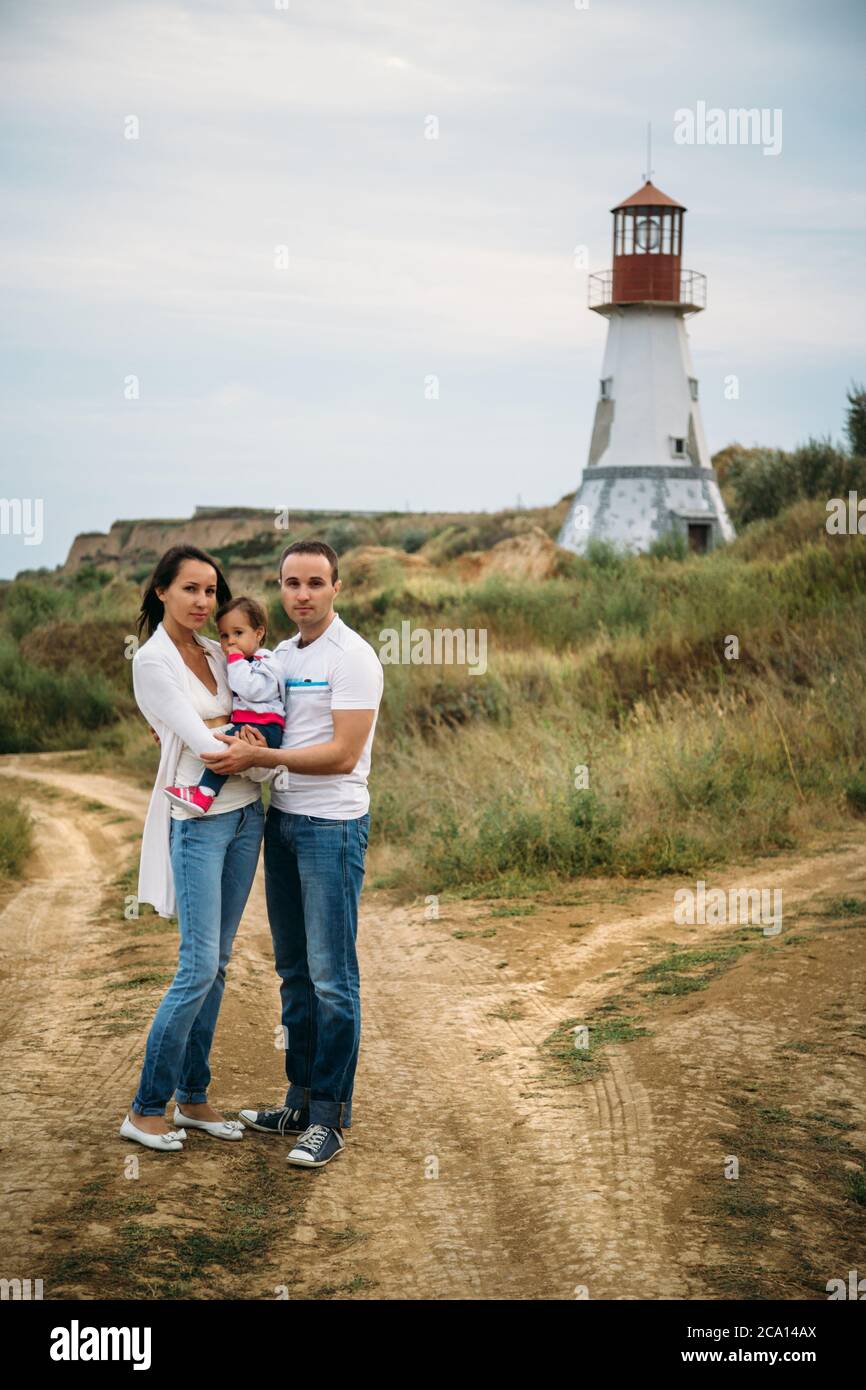 Glückliche junge Familie in weißen T-Shirts und blauen Jeans mit einer kleinen Tochter in der Nähe des weißen Leuchtturms, im Freien Hintergrund mit Möwen am Himmel Stockfoto