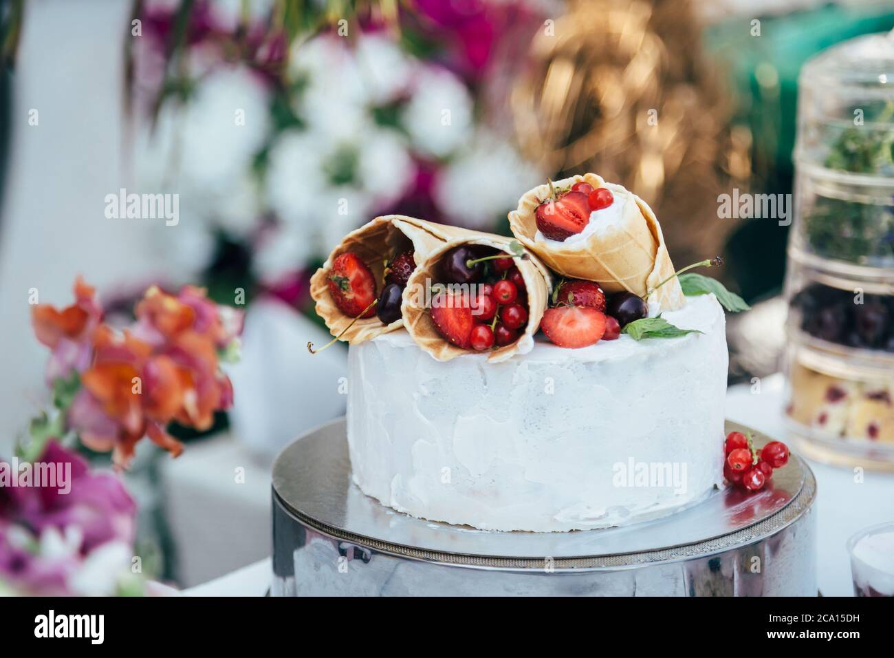 Köstliche weiße cremige Torte mit Waffelkegel gefüllt mit verschiedenen Beeren darauf, umgeben von Blumenschmuck Stockfoto