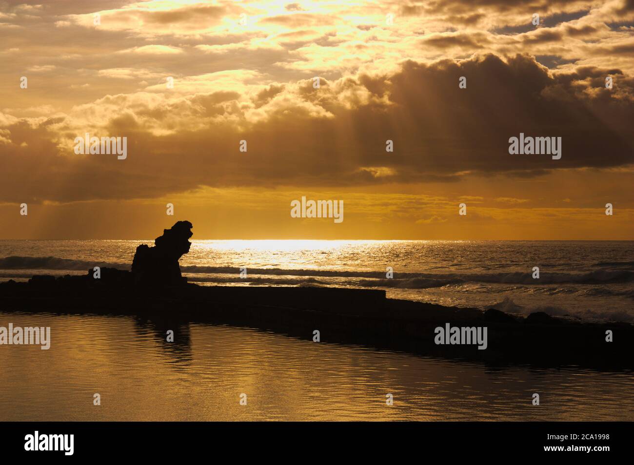 Orangefarbener Sonnenuntergang mit Wolken und Lichtungen von einem Strand mit Felsen im Vordergrund. Romantisches Bild mit Kopierbereich Stockfoto