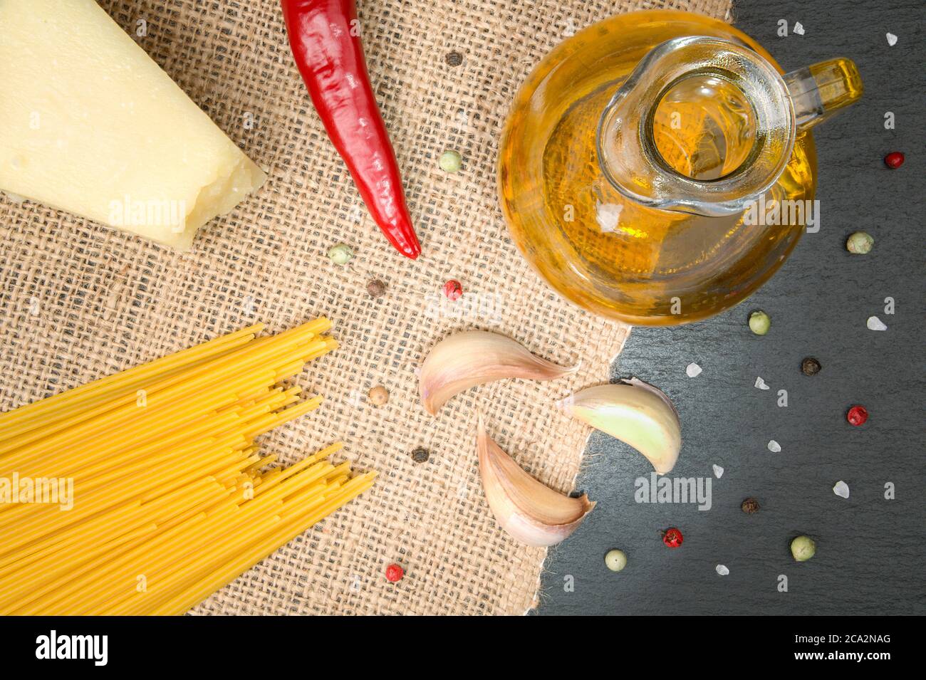 Nahaufnahme der Zutaten von 'Spaghetti Aglio e Olio', teils auf einer Schieferplatte und teils auf einer Sackleine. Mit Spaghetti, einem roten Paprika, einem Krug Olivenöl, ga Stockfoto