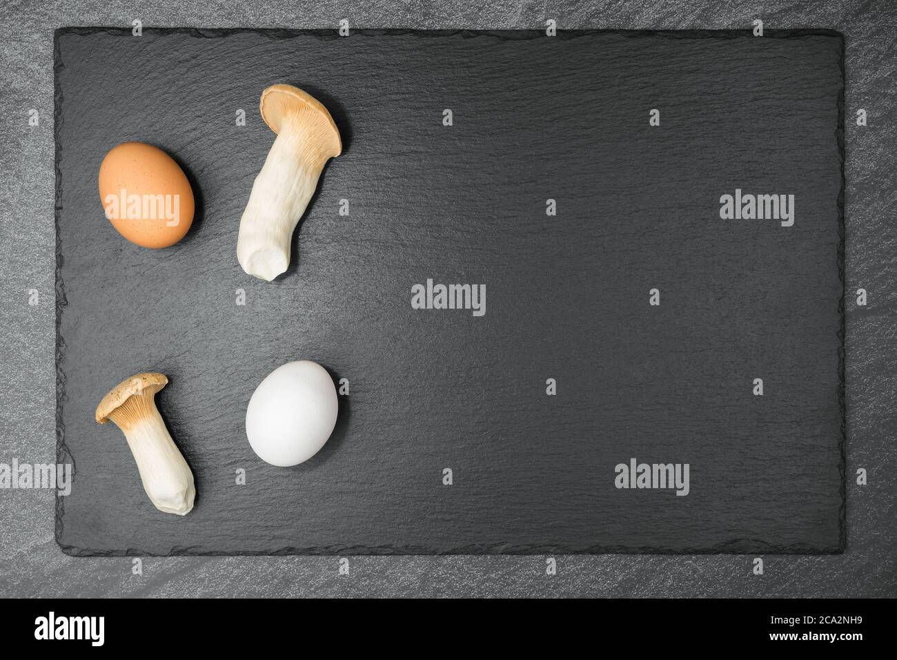 Zwei King Oyster Pilze und ein weißes und ein braunes Ei auf einer Schieferplatte. Mit Copyspace. Flach liegend. Stockfoto