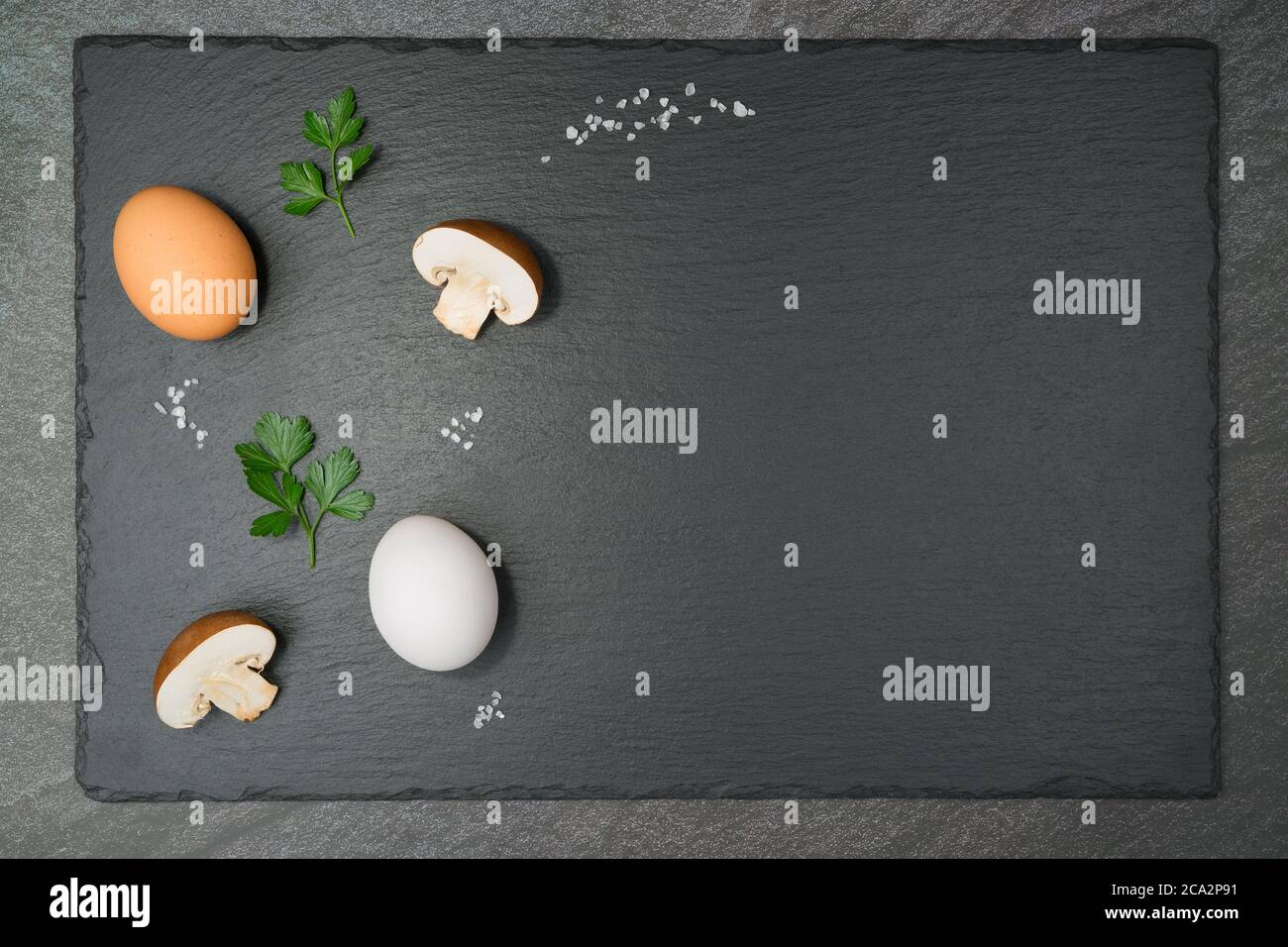Pilz-Omelett-Zutaten mit zwei halbbraunen Pilzen, einem braunen und einem weißen Ei, zwei Petersilienblättern und groben Salzkörnern auf einer Schieferplatte. Mit CO Stockfoto