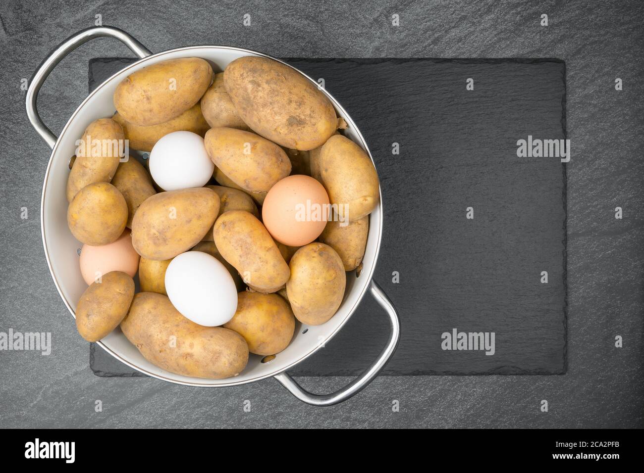 Rohe Kartoffeln und Eier in einem weißen Emailliersieb auf einem Schieferschlag. Flach liegend. Stockfoto