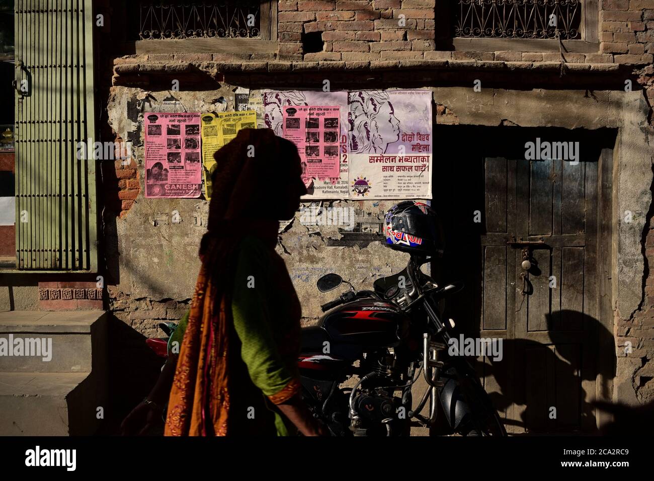 Eine Frau, die auf einer Wohnallee in der Altstadt von Bhaktapur vor einem gemauerten Gebäude mit Werbeplakaten an der Wand läuft. Stockfoto