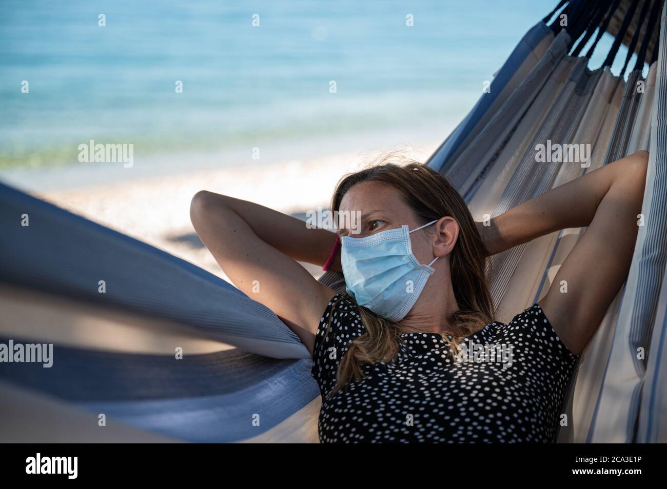 Junge Frau trägt medizinische Schutzmaske in einer Hängematte am schönen Meer entspannen. Stockfoto