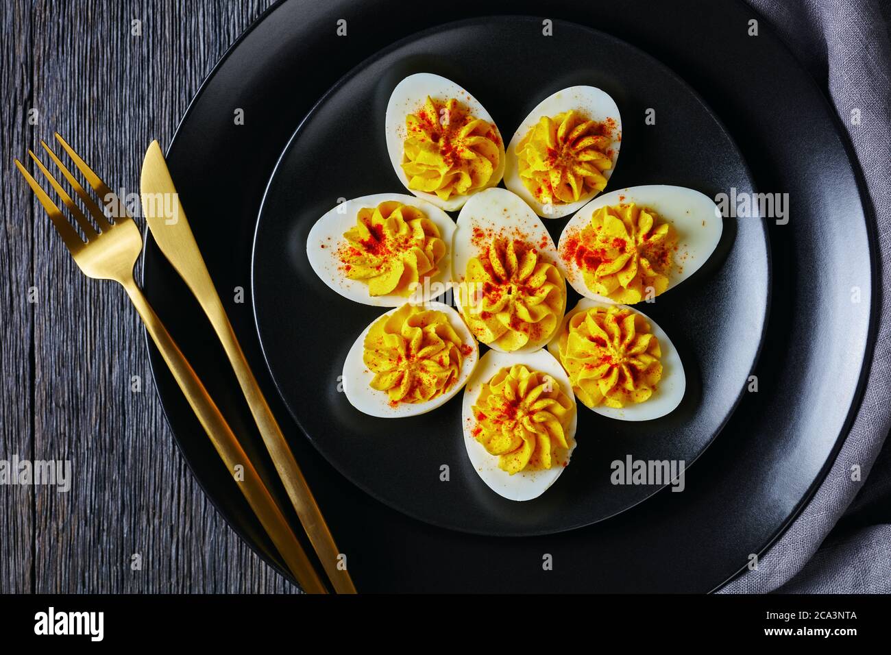 Klassische deviled Eier gefüllt mit Senf, Mayonnaise, Weißessig bestreut mit geräuchertem Paprika auf schwarzem Teller mit goldenem Besteck auf einem dar hölzernen b Stockfoto