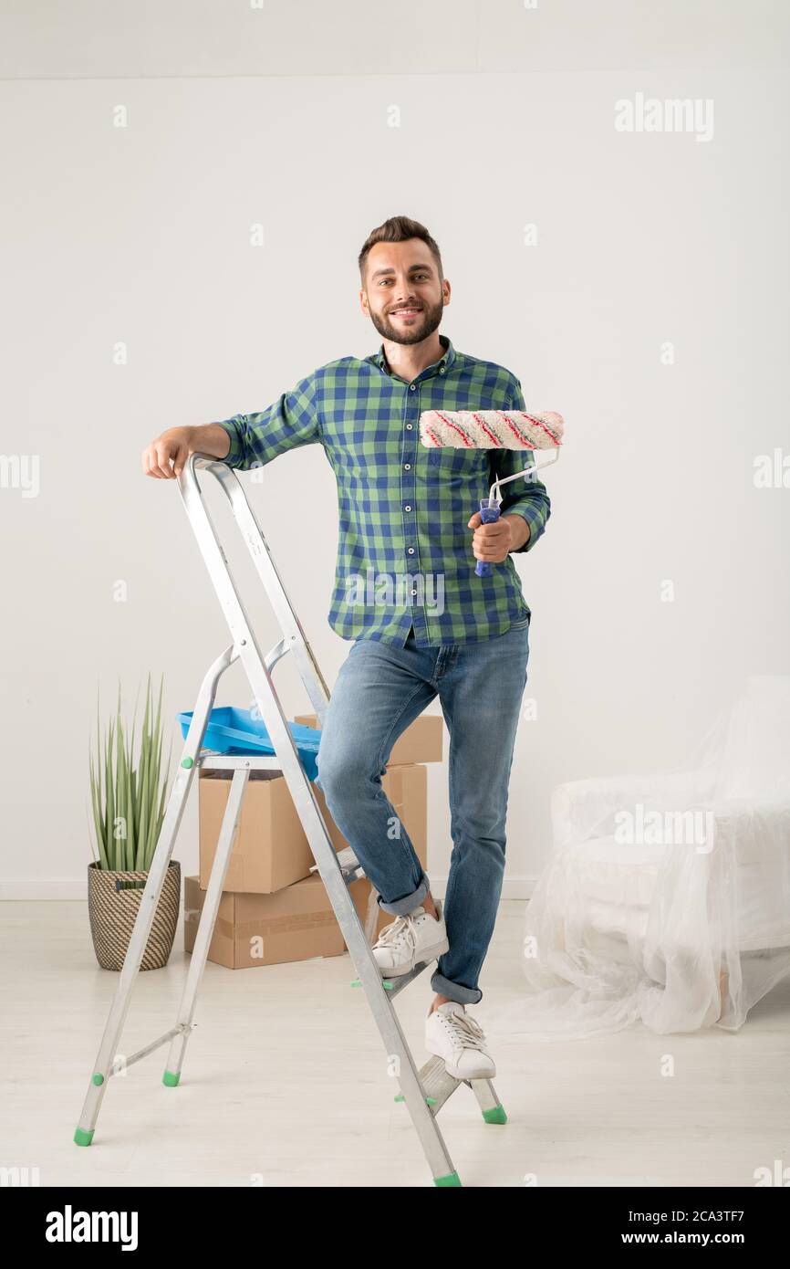 Porträt von fröhlichen jungen bärtigen Mann mit Malerrolle auf der Trittleiter in neue Wohnung mit beweglichen Sachen stehen Stockfoto