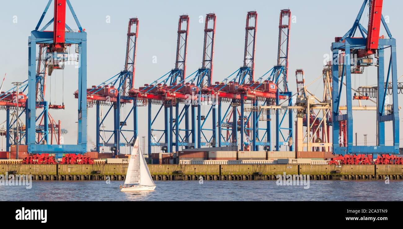 Kleines Segelboot vor einem Containerterminal im Hamburger Hafen. Riesige Hafenkrane im Hintergrund. Stockfoto