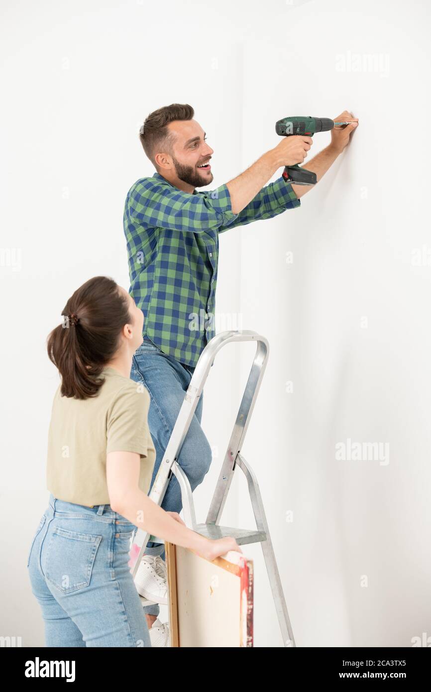 Aufgeregt junge bärtige Mann auf Leiter stehen und drehen Schraube in Wand, während Bild an der Wand mit Frau hängen Stockfoto