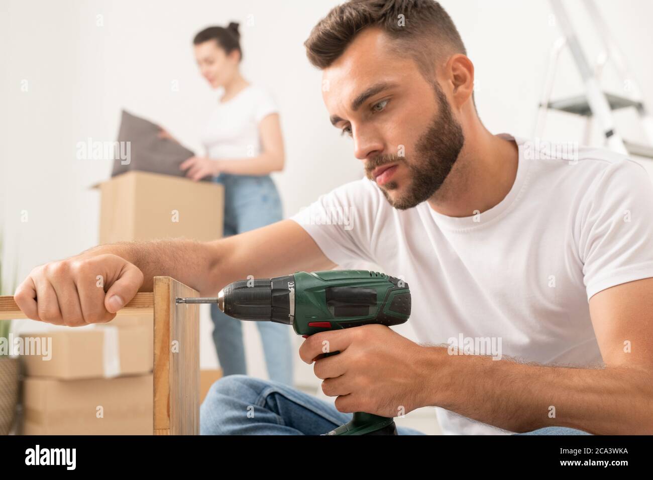 Konzentrierter junger bärtiger Mann, der Möbel mit Akkuschrauber in einer neuen Wohnung zusammenführt Stockfoto