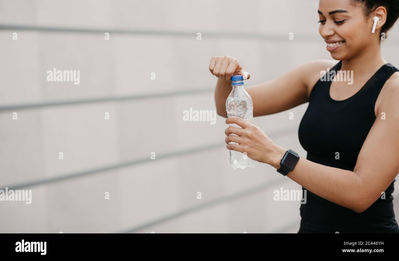 Wasser, gesunde Lebensweise und Bewegung im Freien. Lächelnde afroamerikanische junge Frau mit kabellosem Kopfhörer Öffnung Wasserflasche Stockfoto