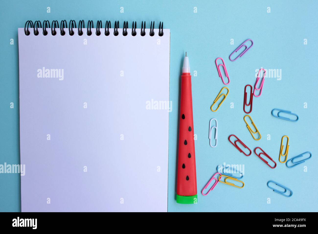 Schulpapier auf blauem Hintergrund. Zurück zur Schule Konzept. Offenes Notizbuch, roter Stift, Radierer, grüner Bleistiftspitzer und bunte Büroklammern Stockfoto