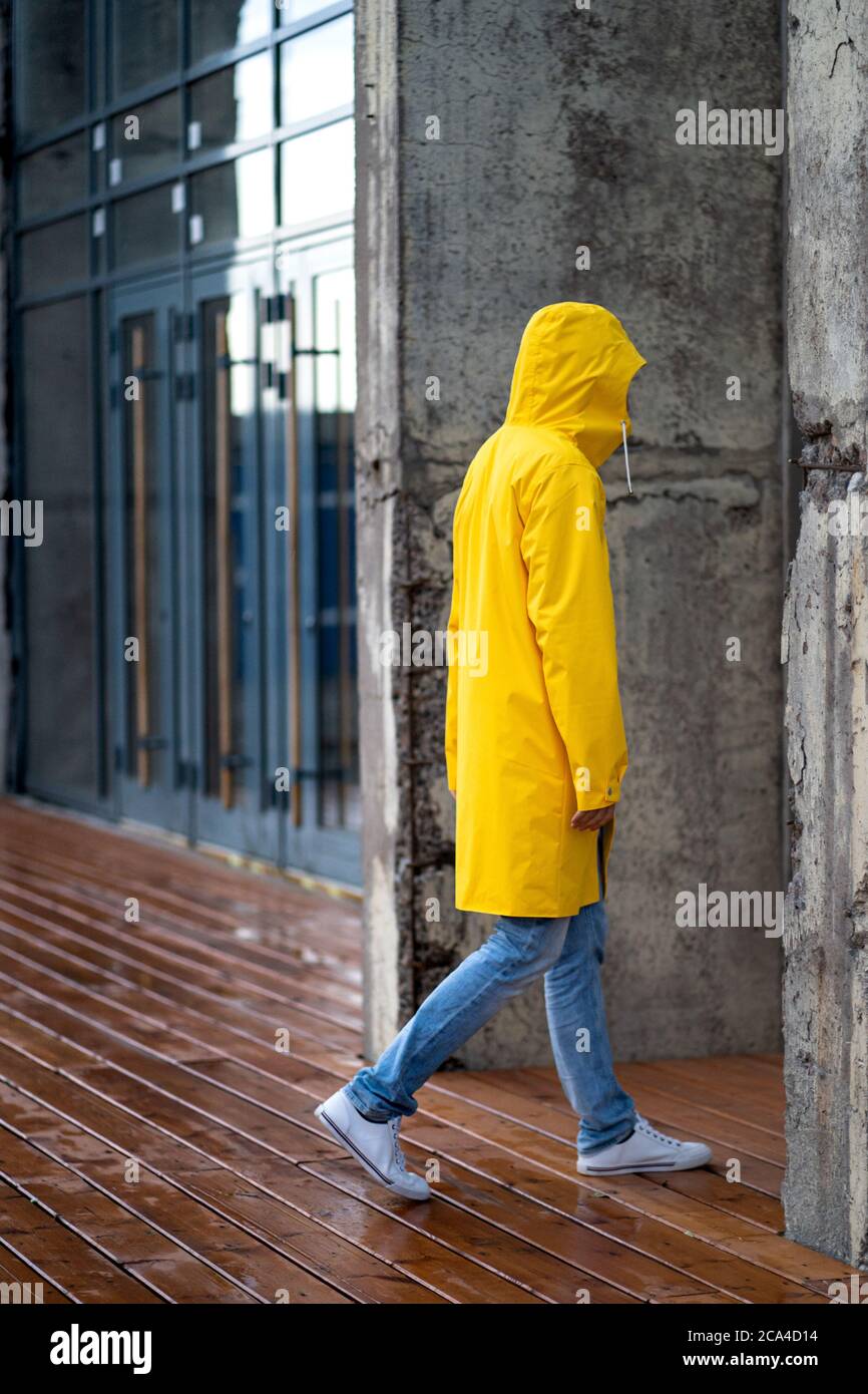 Mann in gelber Regenjacke mit Kapuze auf Spaziergängen bei Regenwetter in der Nähe des Gebäudes auf nassem Holzboden, Seitenansicht. Im Freien. Stockfoto