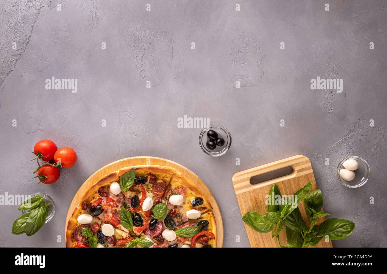 Italienische Pizza mit Schinken, Salami, schwarzen Oliven, Mozzarella-Käse, roten Tomaten und Basilikumblättern auf grauem strukturiertem Hintergrund, umgeben von wenigen Zutaten Stockfoto
