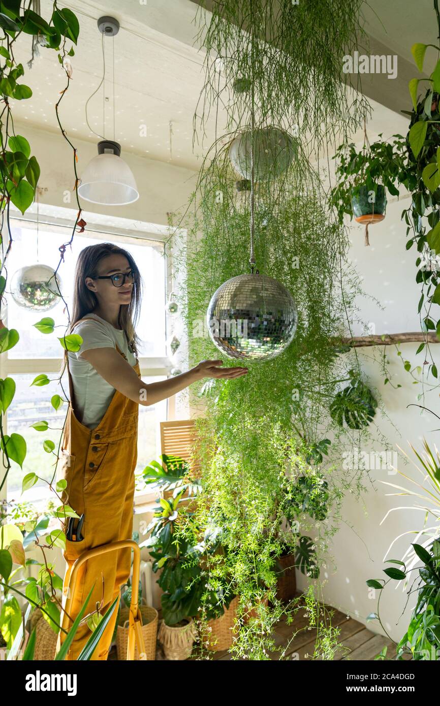 Fröhliche junge Frau Gärtnerin in orange Overalls auf der Trittleiter stehend, berührt die hängende Disco-Ball. Viel Grün zu Hause. Liebe zu Pflanzen. Innengarten Stockfoto