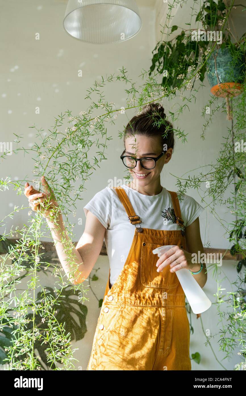 Lächelnde junge Frau Gärtnerin in orange Overalls sprühen üppige Spargelfarn Zimmerpflanze in ihrem Blumengeschäft. Viel Grün zu Hause. Liebe zu Pflanzen. Innen Stockfoto