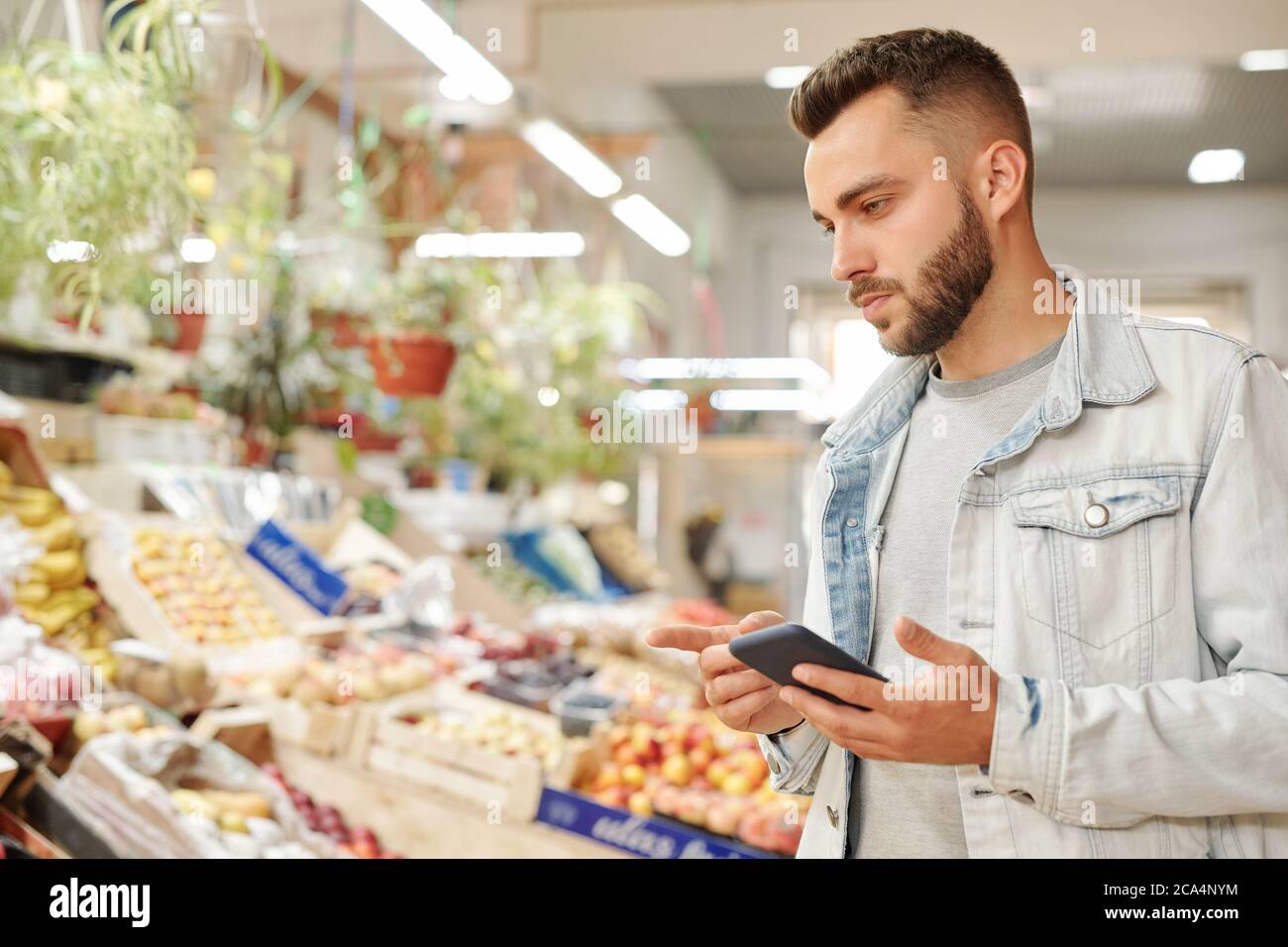 Serious jungen bärtigen Mann in Jacke stehen an der Lebensmitteltheke und überprüfen Lebensmittelliste über Smartphone-App Stockfoto