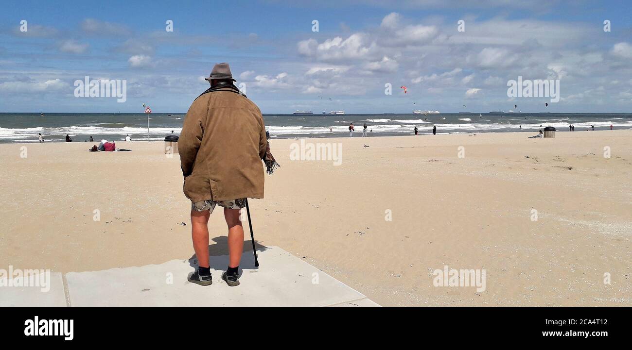 Der alte Mann mit Mantel, kurzer Hose, Hut und Spazierstock blickt auf das Meer an einem Strand in Holland Stockfoto