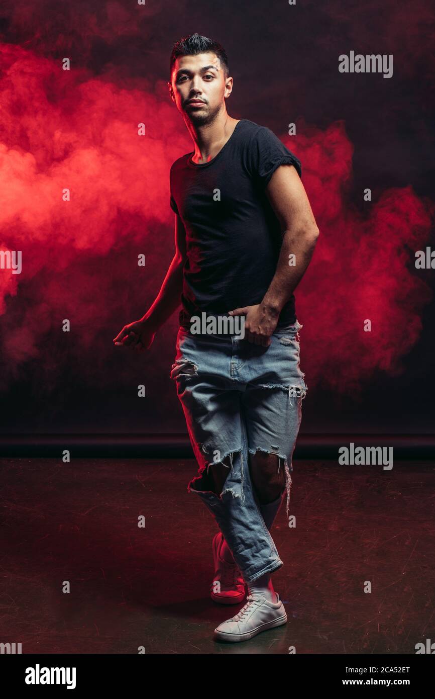 Roter Rauch im Hintergrund, junger Kaukasianer in T-Shirt und Jeans zeigen verschiedene Tanzschritte, Tanzprozess im Studio Stockfoto