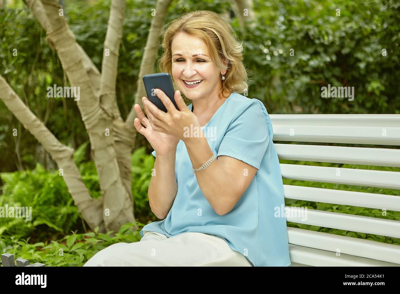 Aktive Rentnerin Attraktive weiße Frau etwa 60 Jahre alt mit Handy in den Händen sitzt auf der Bank im öffentlichen Park. Sie lächelt in ihrem Ga Stockfoto