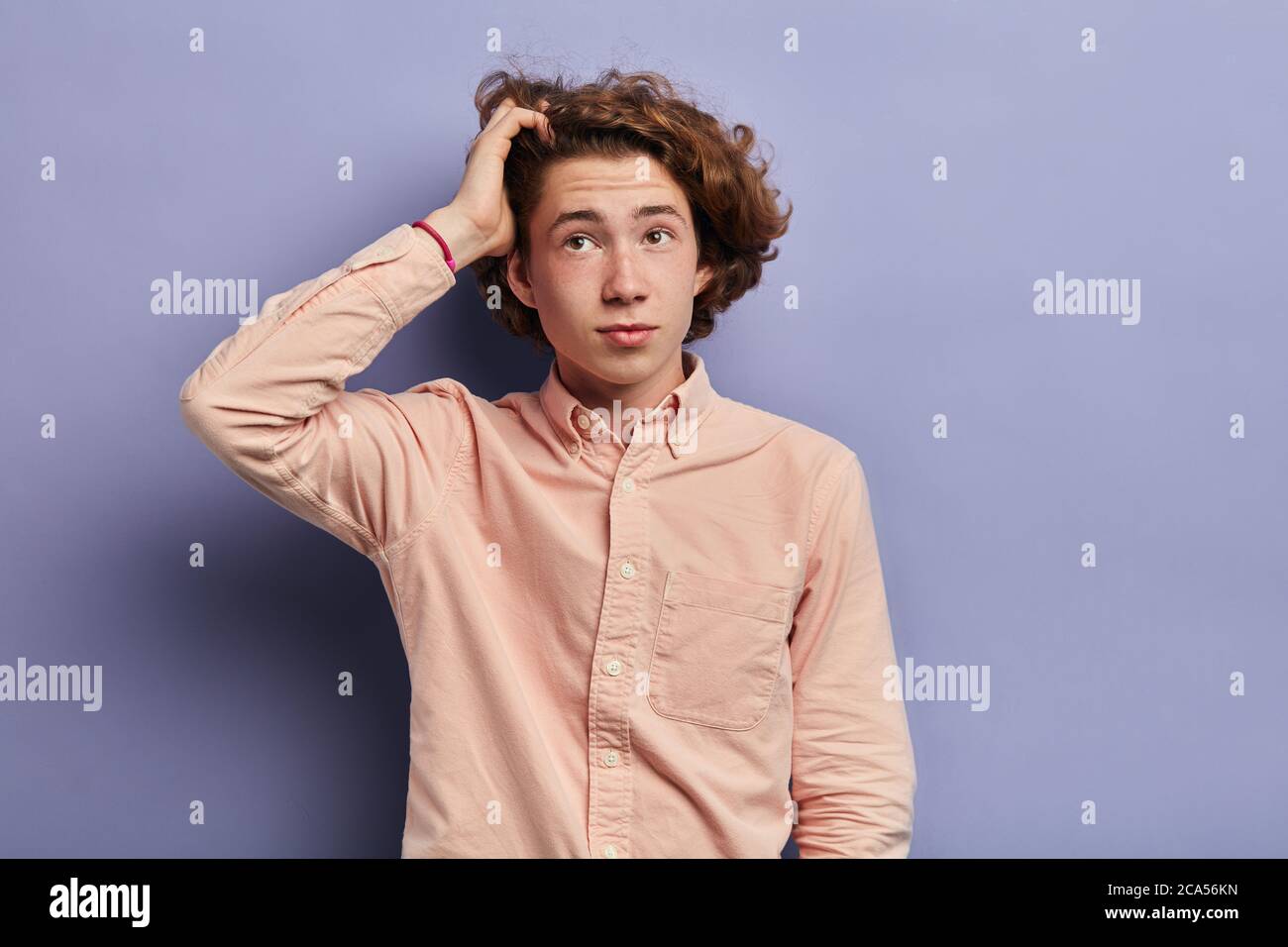 Junge nachdenkliche Mann mit Sommersprossen und welligen langen Haaren, trägt Langarm lässiges helles Hemd, schaut mit verwirrten Ausdruck, behebt lockiges h Stockfoto