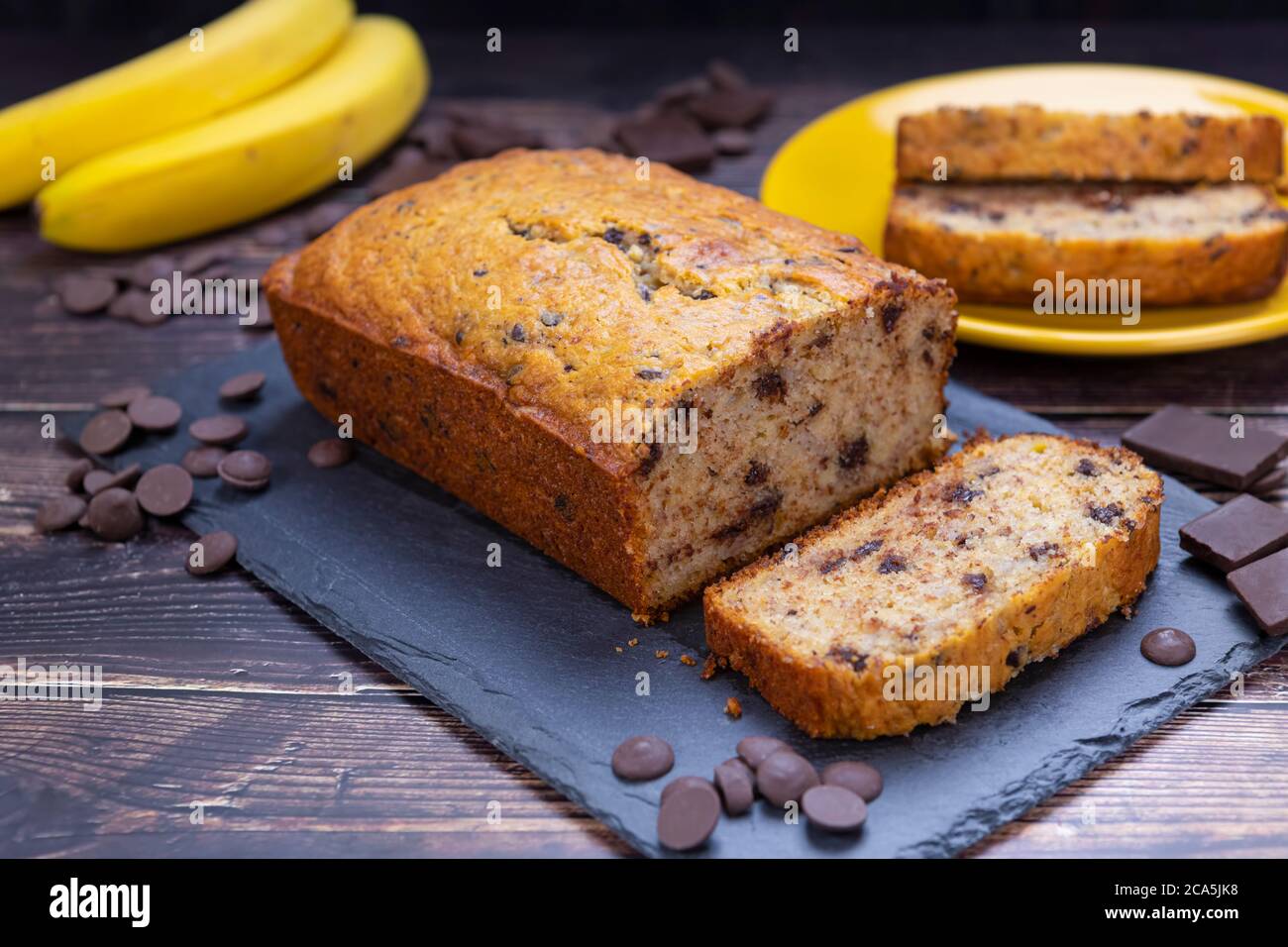 Bananenbrot auf Schneidebrett mit Schokoladenstückchen und frischer Banane im Hintergrund. Stockfoto