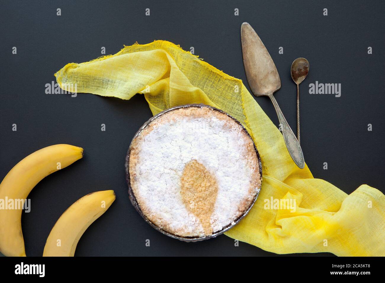 Bananengebundener Kuchen auf schwarzem Hintergrund mit gelber Decke und Retrolöffeln. Hausgemachter Kuchen vor dunkler Kulisse und Löffelschatten Stockfoto