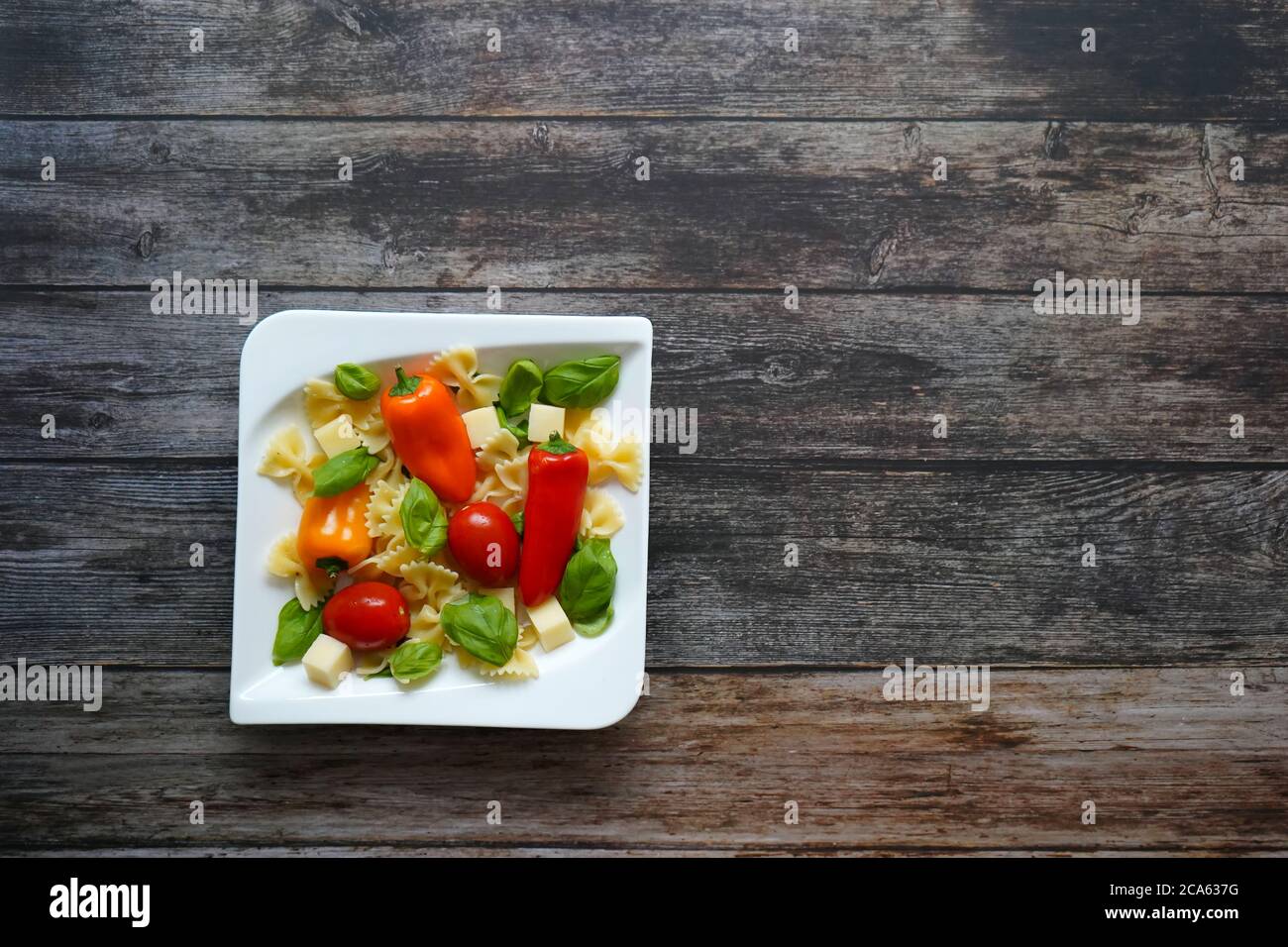 Frischer gesunder Salat mit Nudeln, Tomaten, Paprika, Käsewürfeln und Basilikum auf einem weißen quadratischen Teller. Dunkler Holzhintergrund. Stockfoto