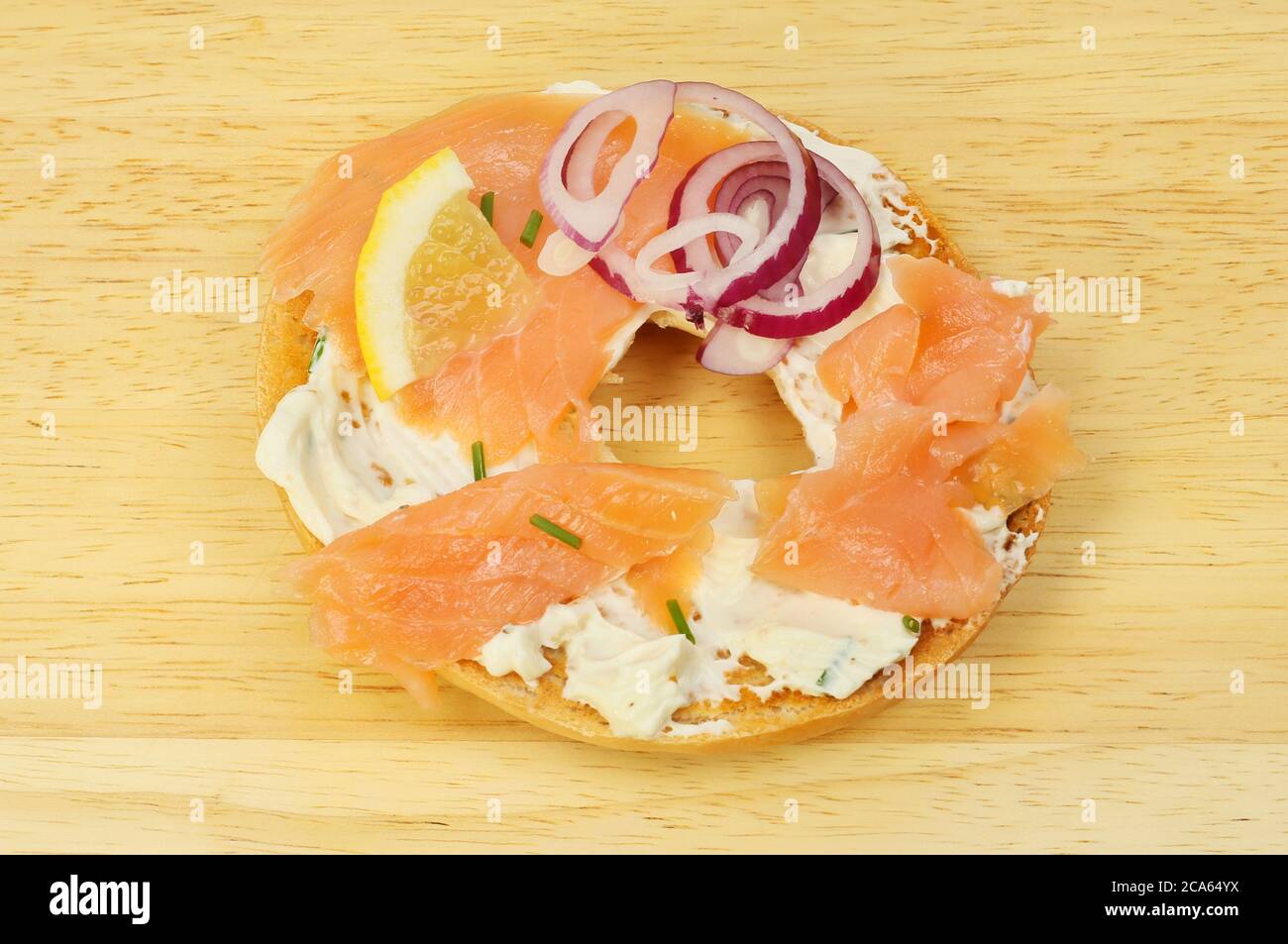 Ein Bagel mit Frischkäse und geräuchertem Lachs, garniert mit Schnittlauch, roten Zwiebeln und Zitronenkeilen auf einem Holzbrett Stockfoto